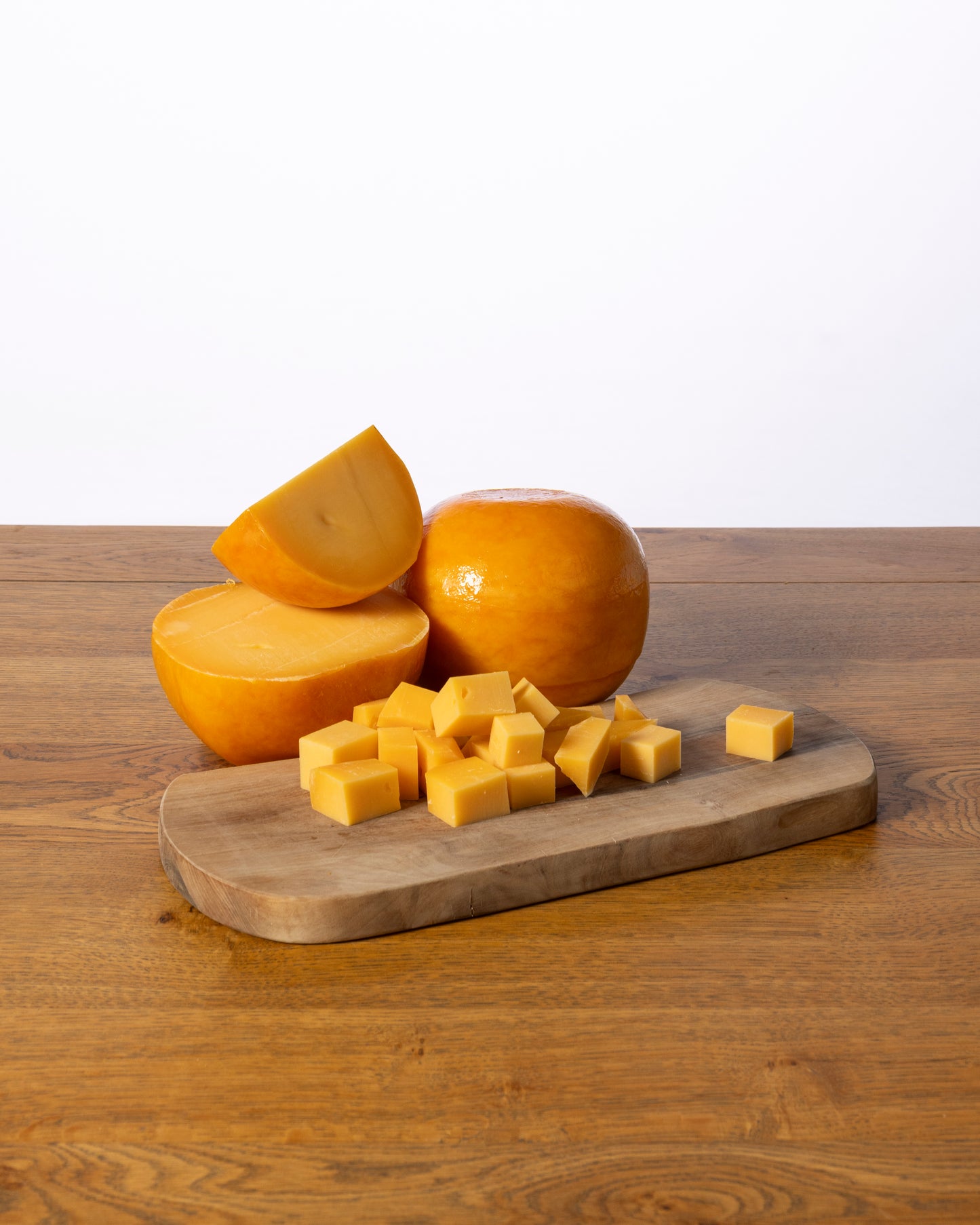 Een bolvormige Edammer 40+ Belegen, in drie stukken gesneden met kleine kaasblokjes ervoor, geplaatst op een houten snijplank.