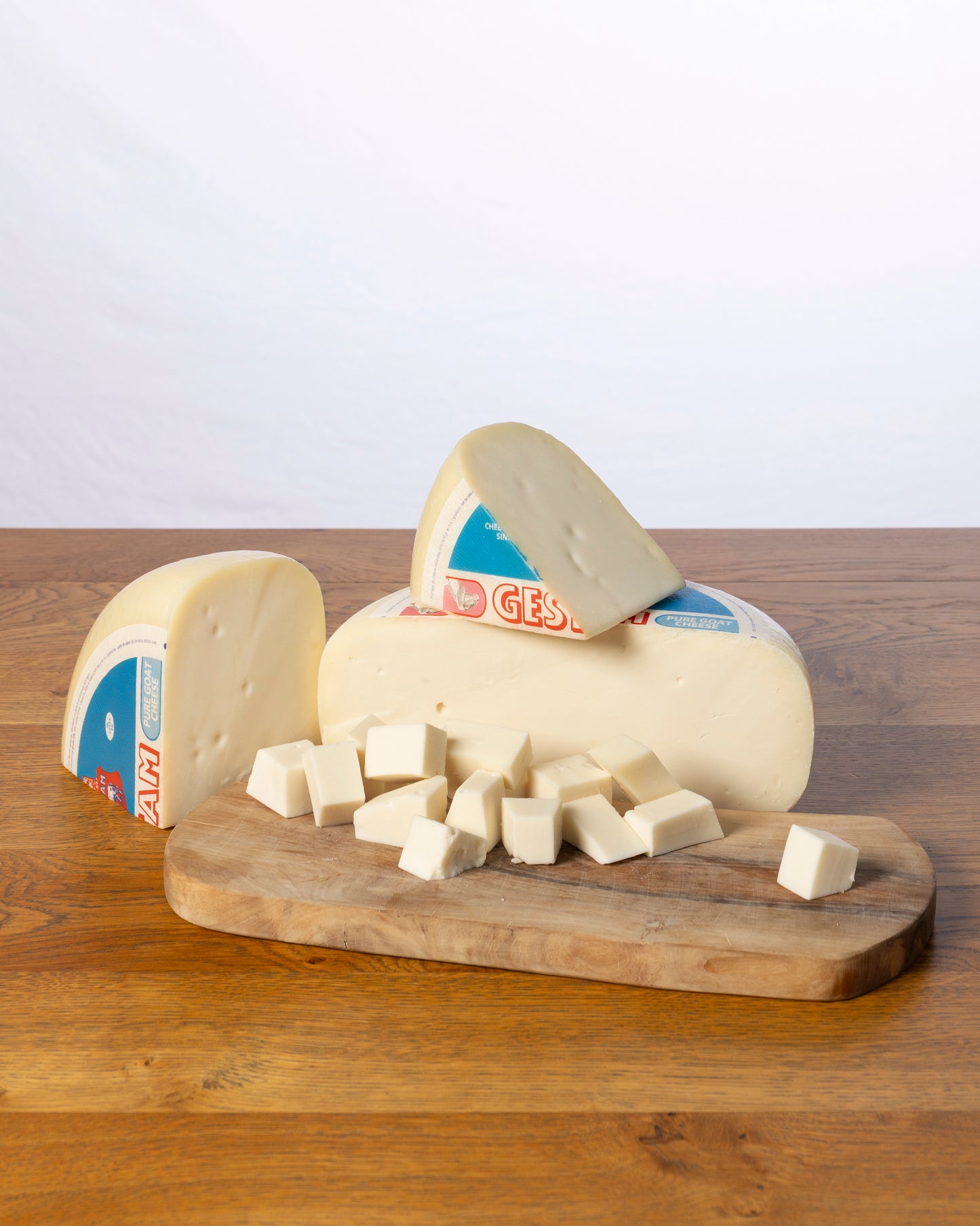 Een wielvormige Geitenkaas 50+ Jong, in drie stukken gesneden met kleine kaasblokjes ervoor, geplaatst op een houten snijplank.