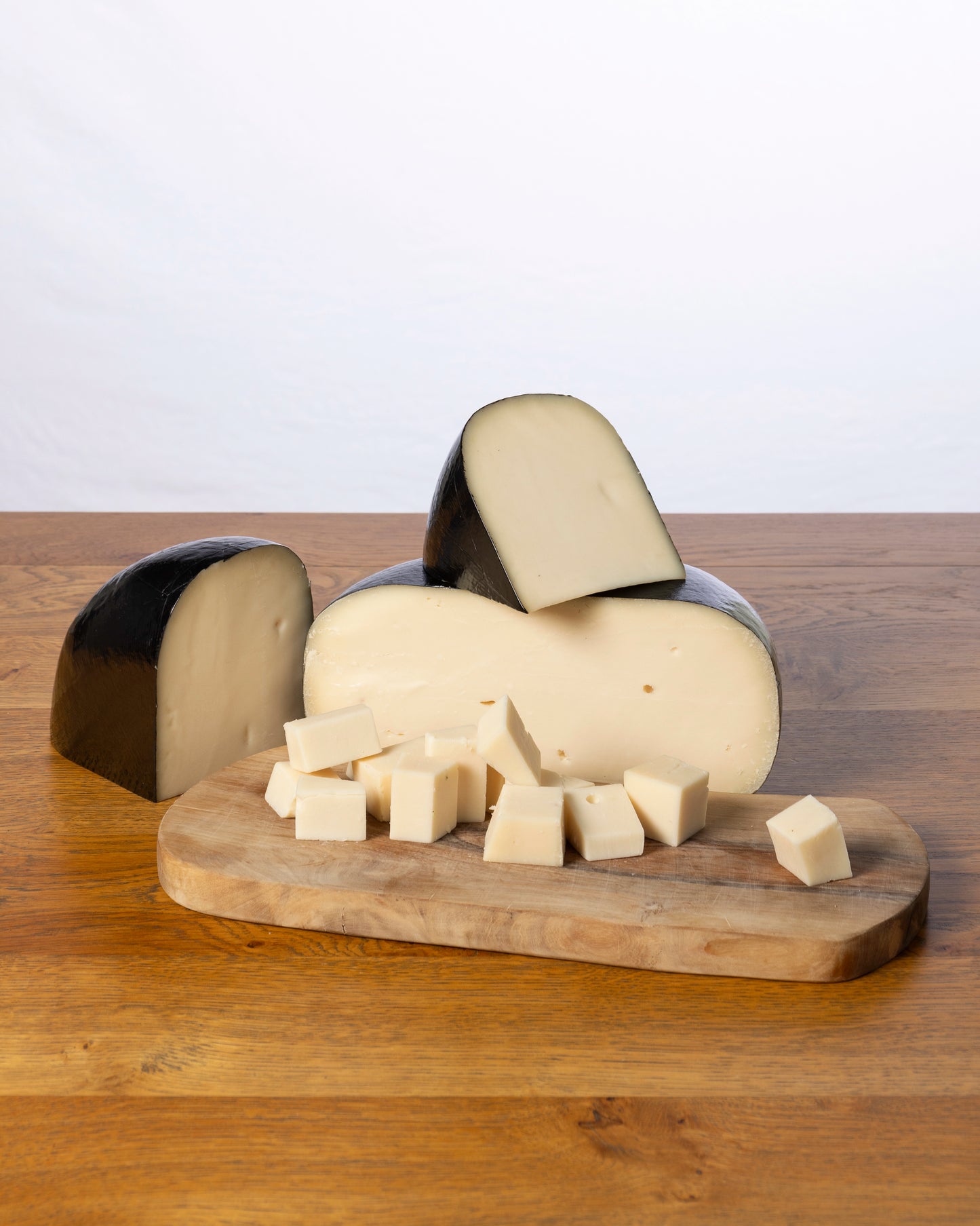 Een wielvormige Geitenkaas 50+ Oud, in drie stukken gesneden met kleine kaasblokjes ervoor, geplaatst op een houten snijplank.