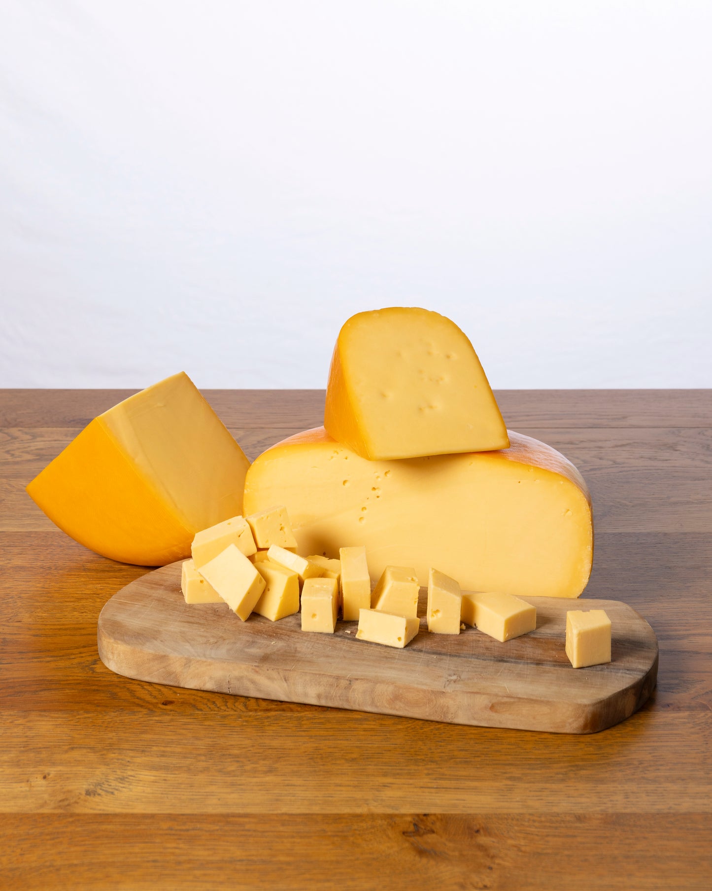Een wielvormige Goudse 50+ Jong Belegen, in drie stukken gesneden met kleine kaasblokjes ervoor, geplaatst op een houten snijplank.