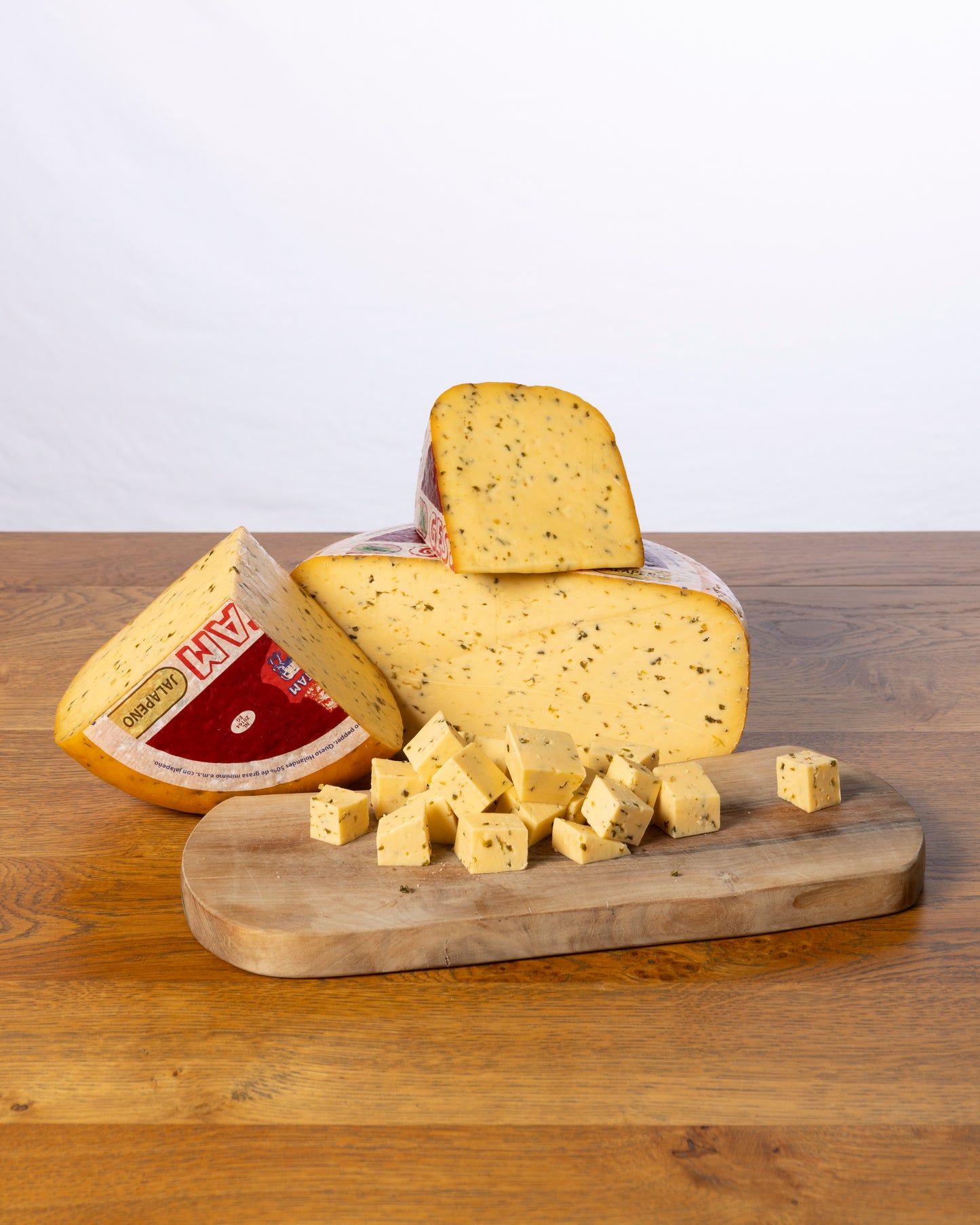 Een wielvormige Jalapeño 50+ Jong kaas, in drie stukken gesneden met kleine kaasblokjes ervoor, geplaatst op een houten snijplank.